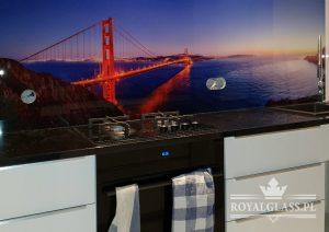 Szklany panel kuchenny z podświetlanym motywem mostu Golden Gate w San Francisco, zamontowany nad płytą grzewczą i piekarnikiem w nowoczesnej kuchni z białymi szafkami i czarnym blatem.