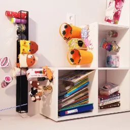 Kącik dziecięcy z białym regałem wypełnionym książkami, zabawkami i dekoracjami, w tym ręcznie robionymi ozdobami i wiatraczkiem na ścianie.