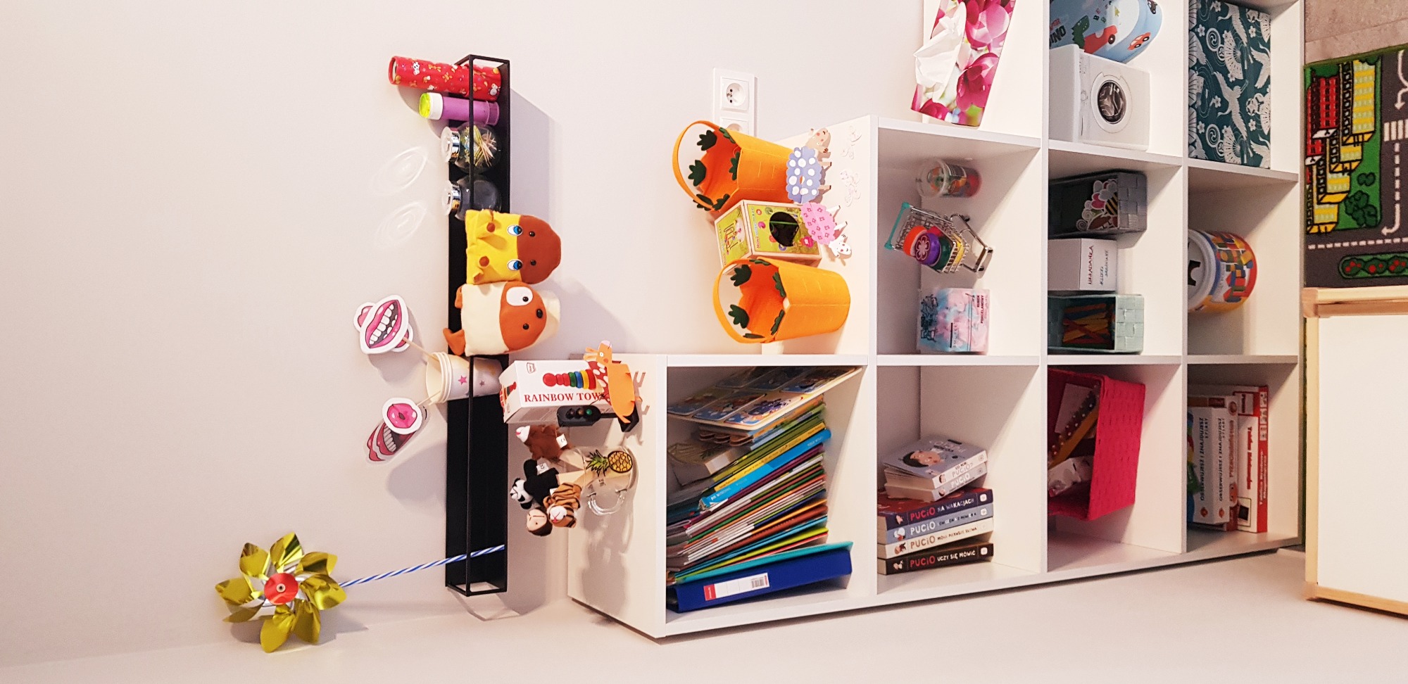 Kącik dziecięcy z białym regałem wypełnionym książkami, zabawkami i dekoracjami, w tym ręcznie robionymi ozdobami i wiatraczkiem na ścianie.