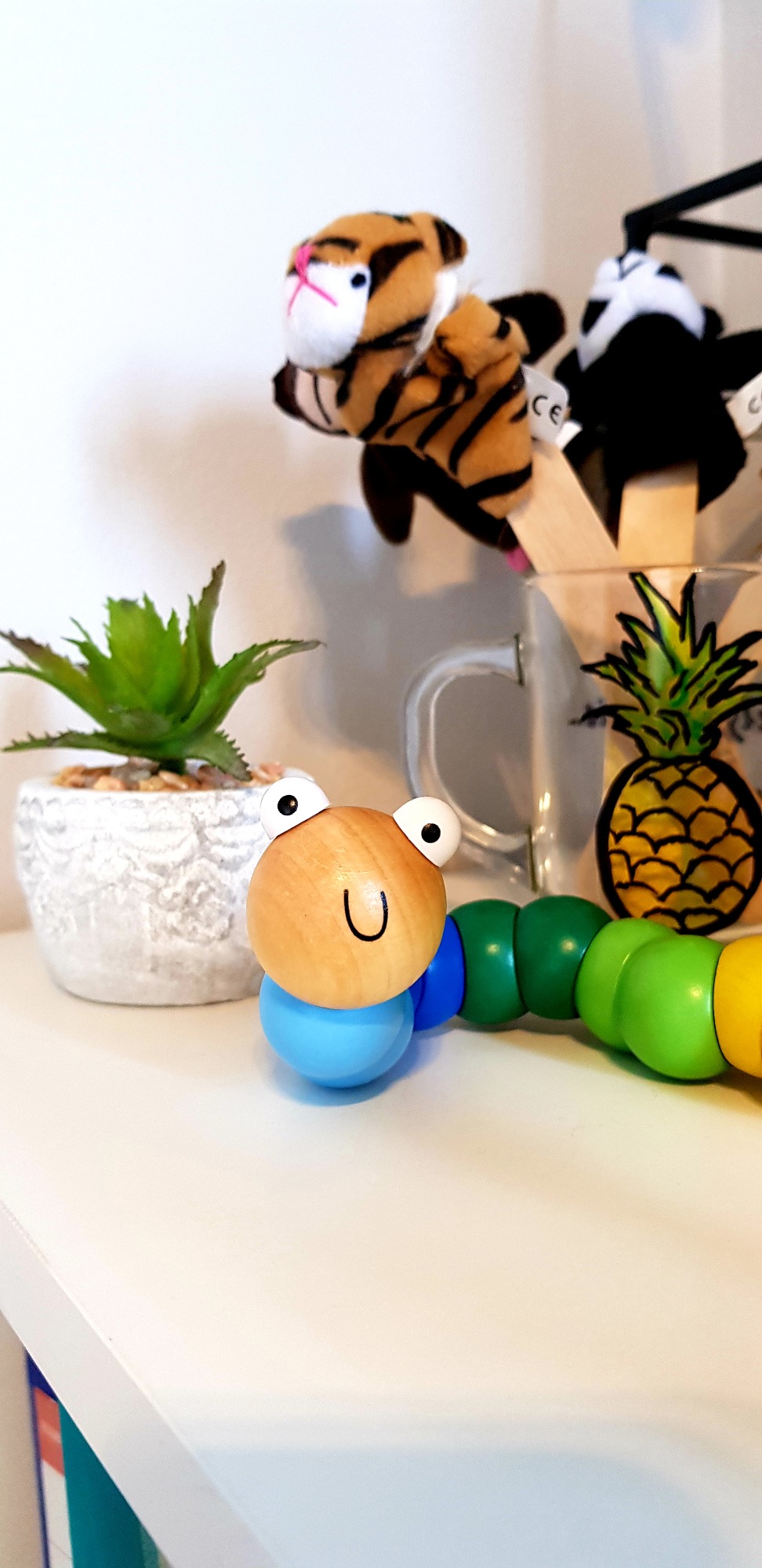Drewniany, kolorowy robaczek z uśmiechniętą buzią i wyłupiastymi oczami na białej półce, obok doniczki z sukulentem i kubka z ananasem, w tle pluszowe pacynki na patyku.