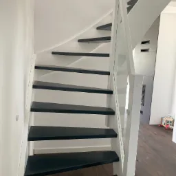 Nowoczesne schody z czarnymi stopniami i białymi elementami konstrukcyjnymi, z przeszkloną balustradą, widok z dołu na górę.