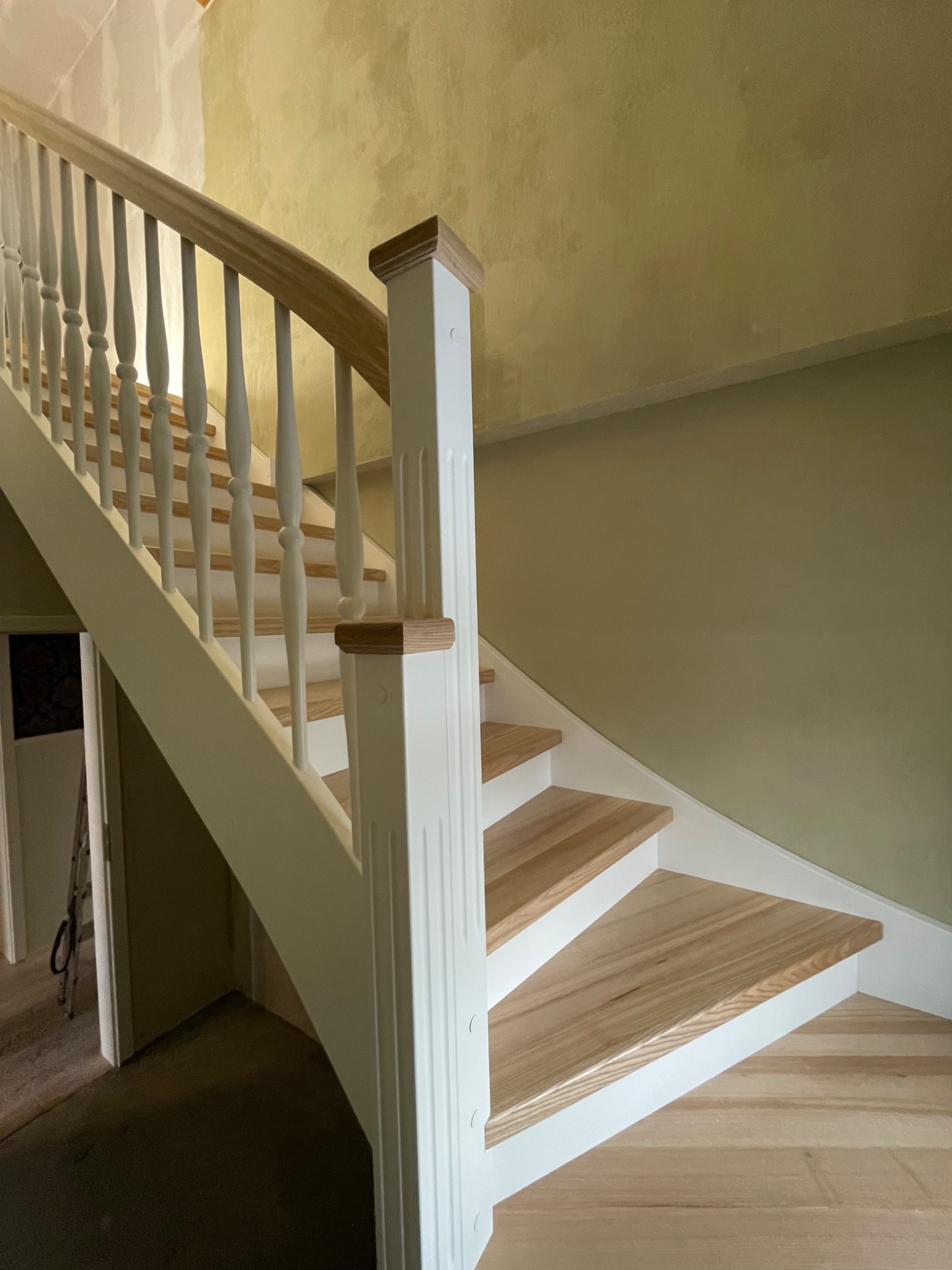 Klasyczne schody drewniane z białymi balustradami i drewnianymi stopniami, widoczne niedokończone malowanie ściany w tle.