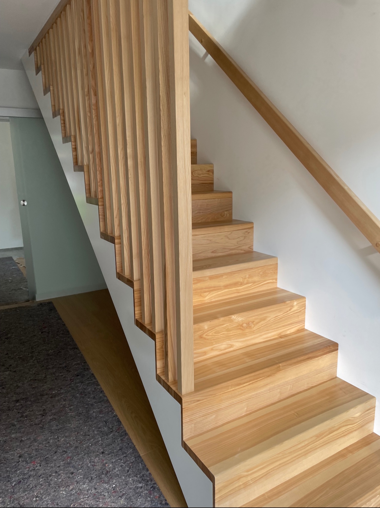 Jasne, drewniane schody z pionowymi szczeblami balustrady i poręczą w minimalistycznym wnętrzu. Widoczne zabezpieczenie podłogi sugeruje trwający montaż.