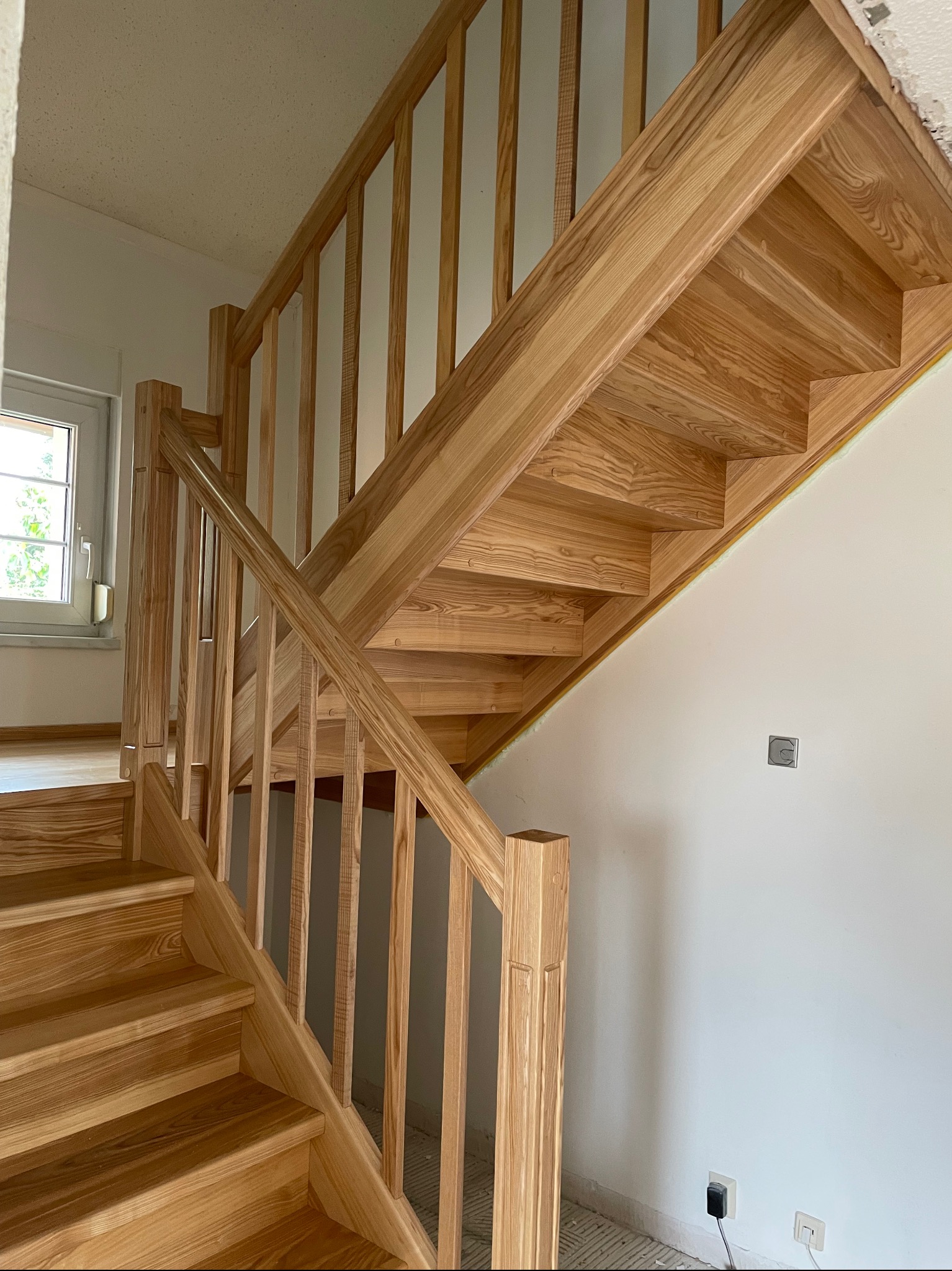 Jasne, drewniane schody z balustradą w trakcie montażu, widoczne niedokończone wykończenia ścian i podłóg, naturalne oświetlenie wpadające przez okno.