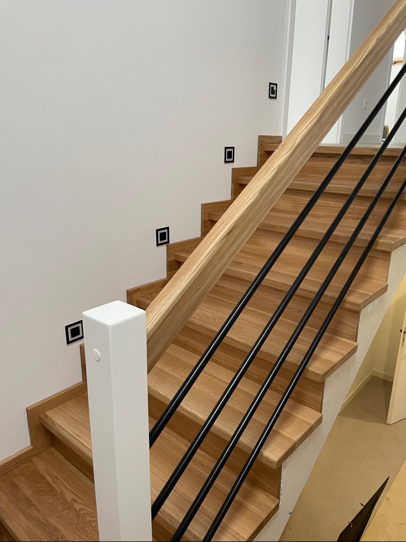 Jasne, drewniane stopnie z czarnymi, metalowymi prętami balustrady i drewnianą poręczą, oświetlone małymi, kwadratowymi lampami ściennymi.