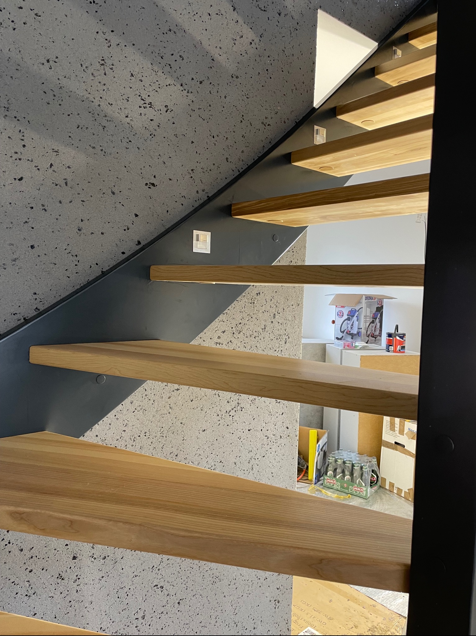 Nowoczesne schody z drewnianymi stopniami i szarą, betonową ścianą. Widoczne elementy konstrukcyjne i kartony w tle.