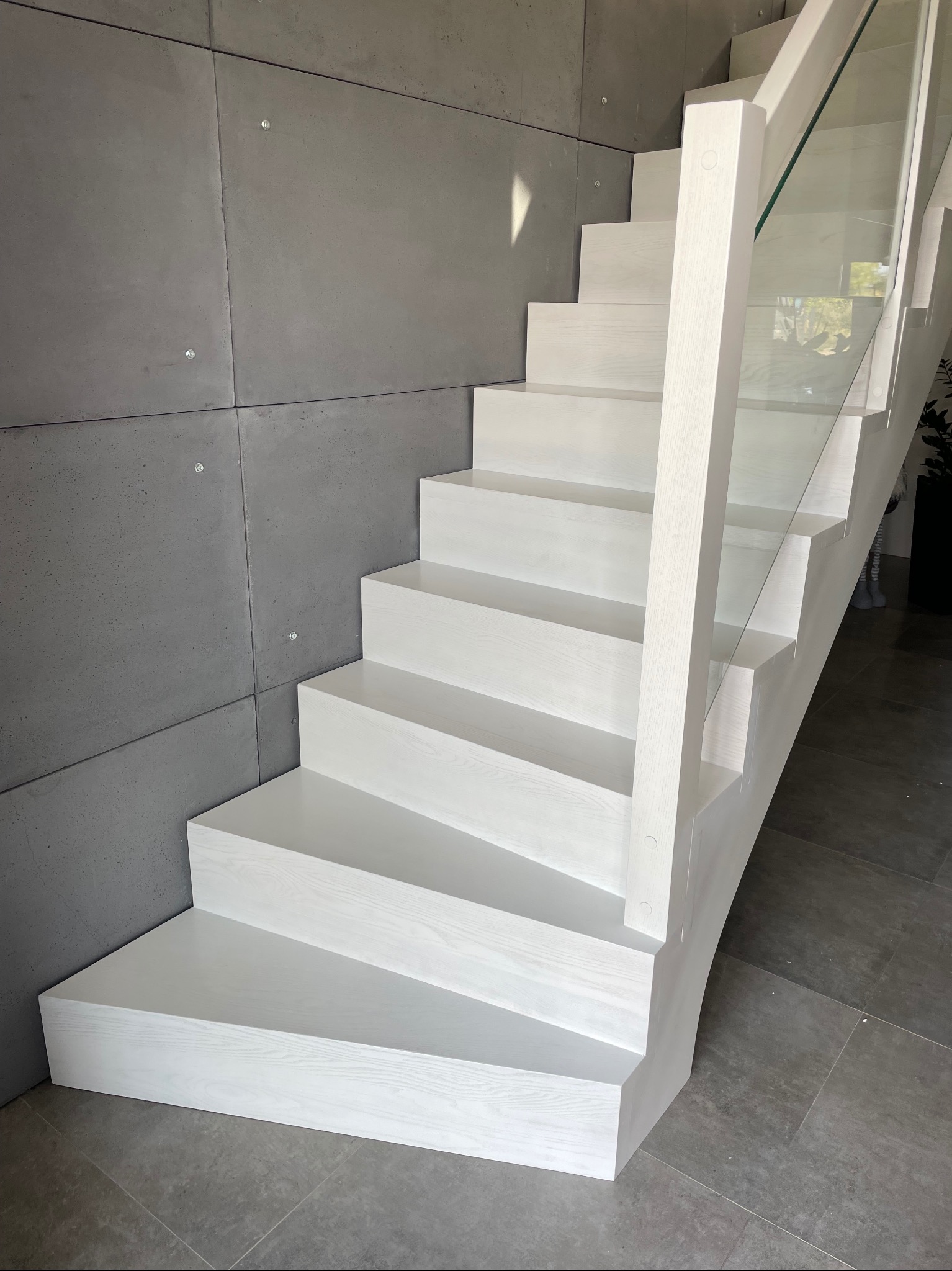 Białe, drewniane schody z przeszkloną balustradą, przylegające do ściany wykończonej betonowymi panelami, na podłodze szare płytki.
