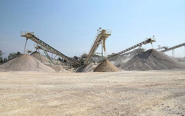 Szeroki kadr na kopalnię piasku i żwiru z taśmociągami i usypiskami różnych frakcji, w tle drzewa i błękitne niebo.