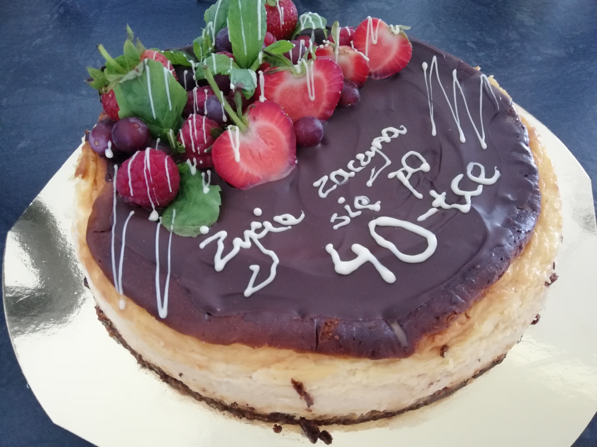 Sernik z polewą czekoladową, dekorowany świeżymi truskawkami, malinami, śliwkami i listkami mięty, z okolicznościowym napisem wykonanym białą polewą.