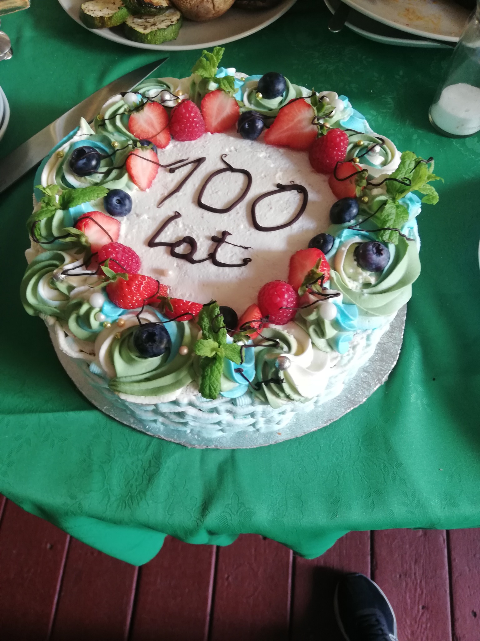 Tort urodzinowy z kremem w odcieniach bieli, zieleni i błękitu, udekorowany świeżymi truskawkami, malinami, borówkami i listkami mięty, z napisem '100 lat' wykonanym czekoladą, leżący na zielonym...