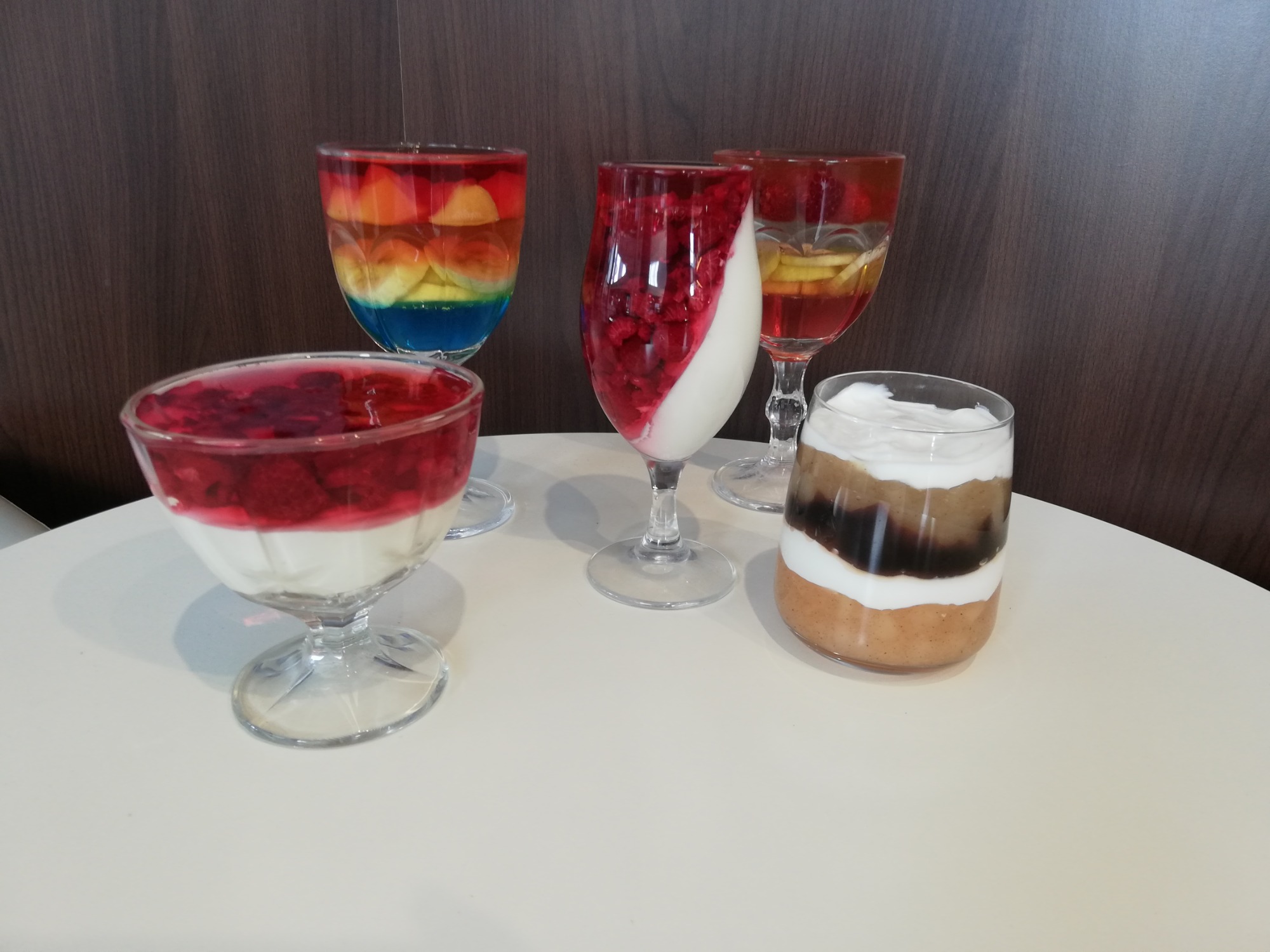 Cztery desery w szklanych naczyniach: tęczowy deser z owocami, deser z malinami i śmietaną, deser z kawałkami jabłek w galaretce i deser z kremem i sosem karmelowym.