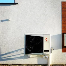 Biała pompa ciepła Rotenso miniRVRF V5 zamontowana na elewacji budynku z widocznym fragmentem drewnianej okładziny, oświetlona popołudniowym słońcem rzucającym cień.