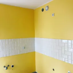 Żółta ściana z poziomym pasem białych, błyszczących płytek ułożonych w jodełkę, widoczne gniazdka elektryczne i otwory wentylacyjne.
