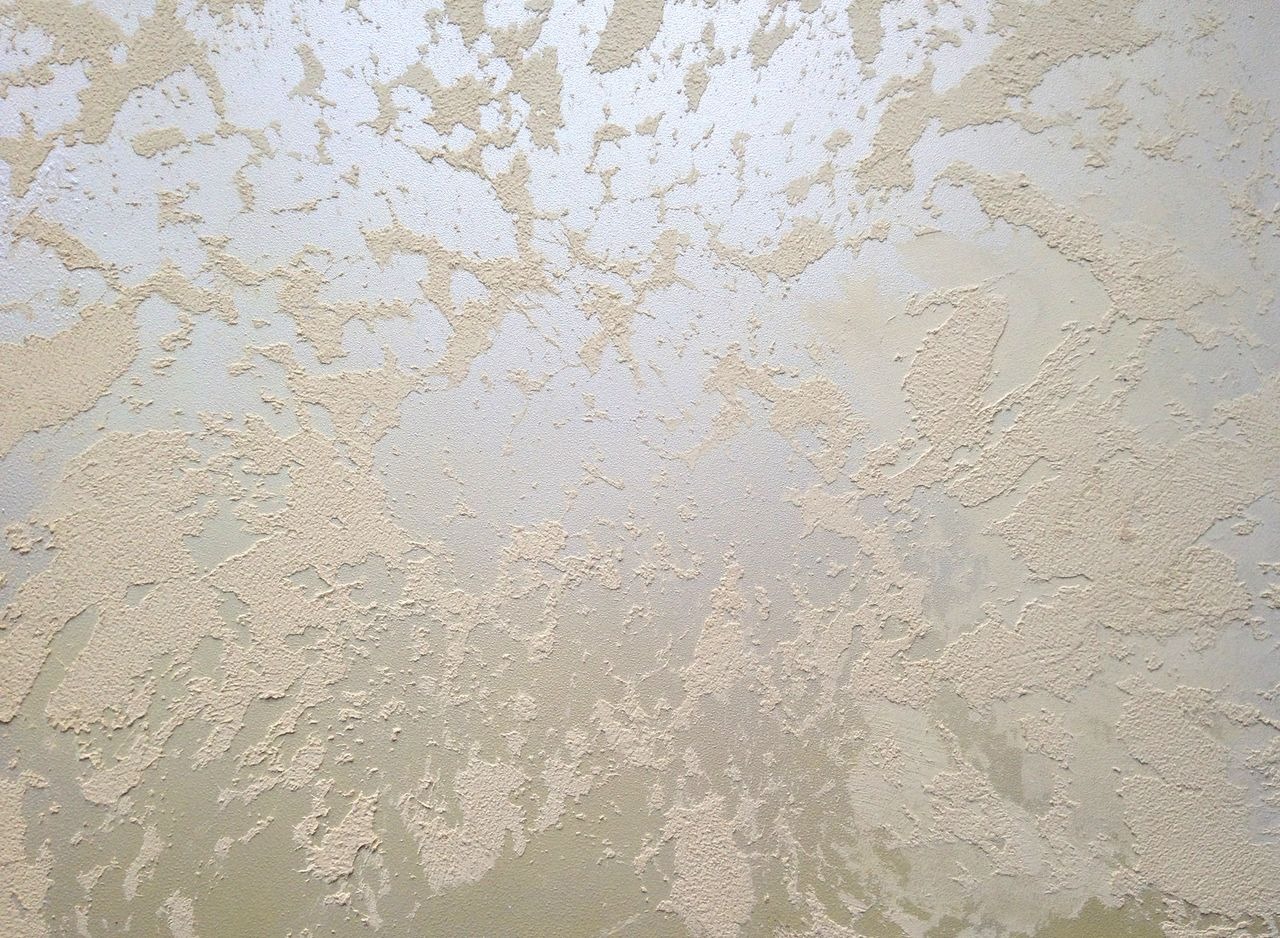 Beżowa ściana z dekoracyjnym, nieregularnym wzorem strukturalnym, imitującym naturalne spękania i nierówności.