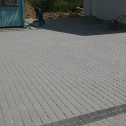 Szeroki podjazd z szarej kostki brukowej z ciemnoszarym obramowaniem, widoczny pracownik w niebieskim kombinezonie i ubijarka w tle.