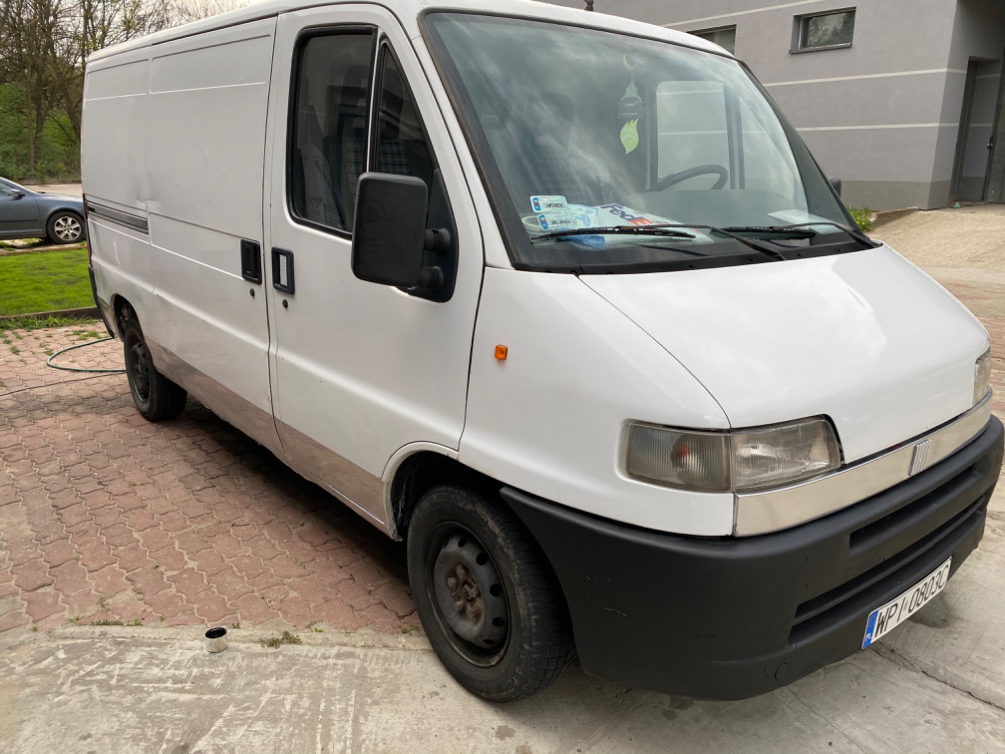 Biały samochód dostawczy Fiat Ducato zaparkowany na czerwonej kostce brukowej, widoczny z przodu pod lekkim kątem, z kubkiem po farbie leżącym obok przedniego koła.