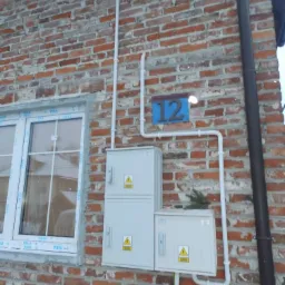 Zewnętrzna skrzynka elektryczna zamontowana na ceglanej ścianie budynku, z widocznymi przewodami w białych rurkach PCV, numer domu 12 na niebieskiej tabliczce, obok okna z białymi ramami.