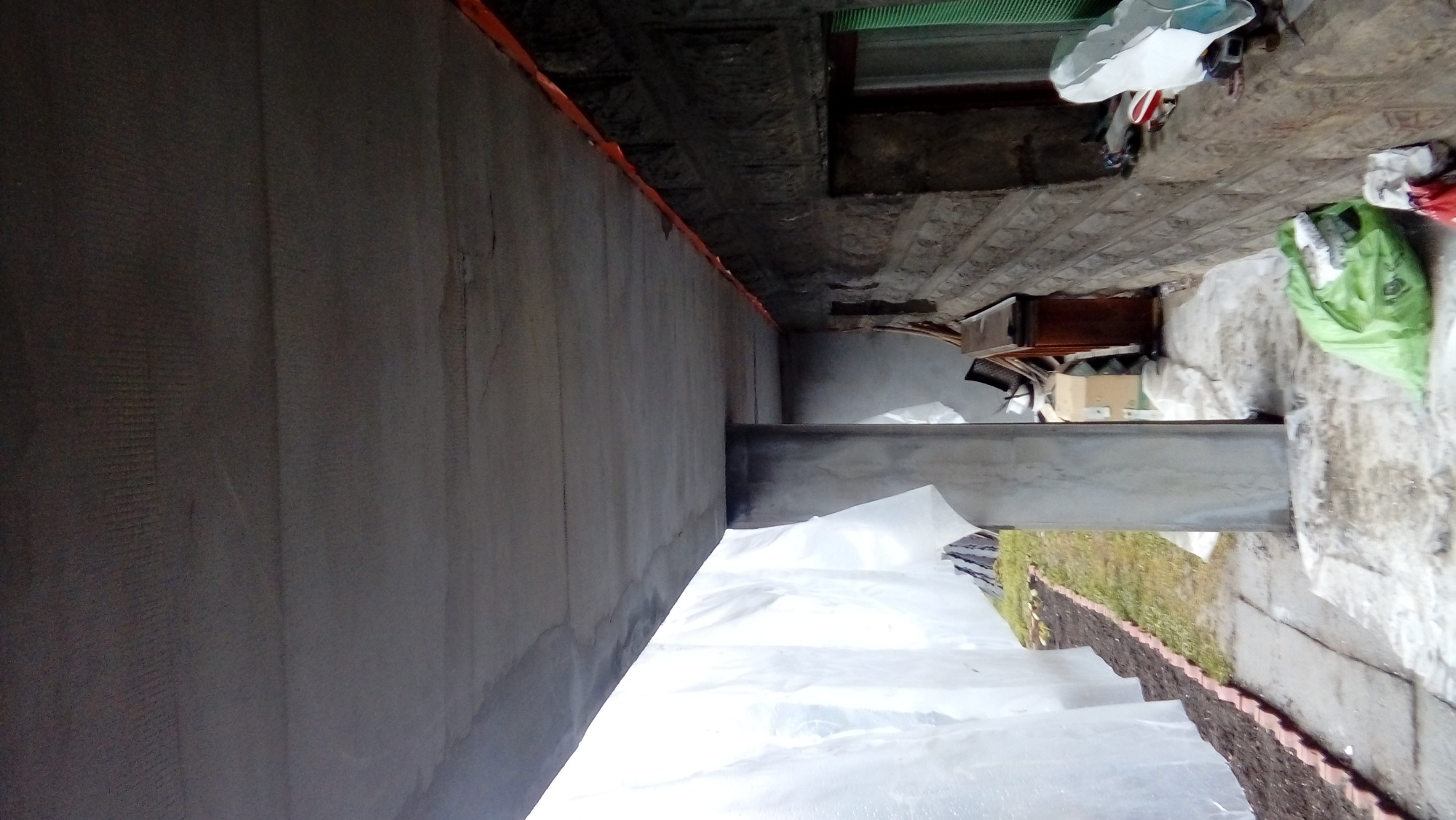 Surowa ściana z szarym tynkiem, widoczne ślady po łatach, zabezpieczona folią malarską, w tle widoczne narzędzia i materiały budowlane.