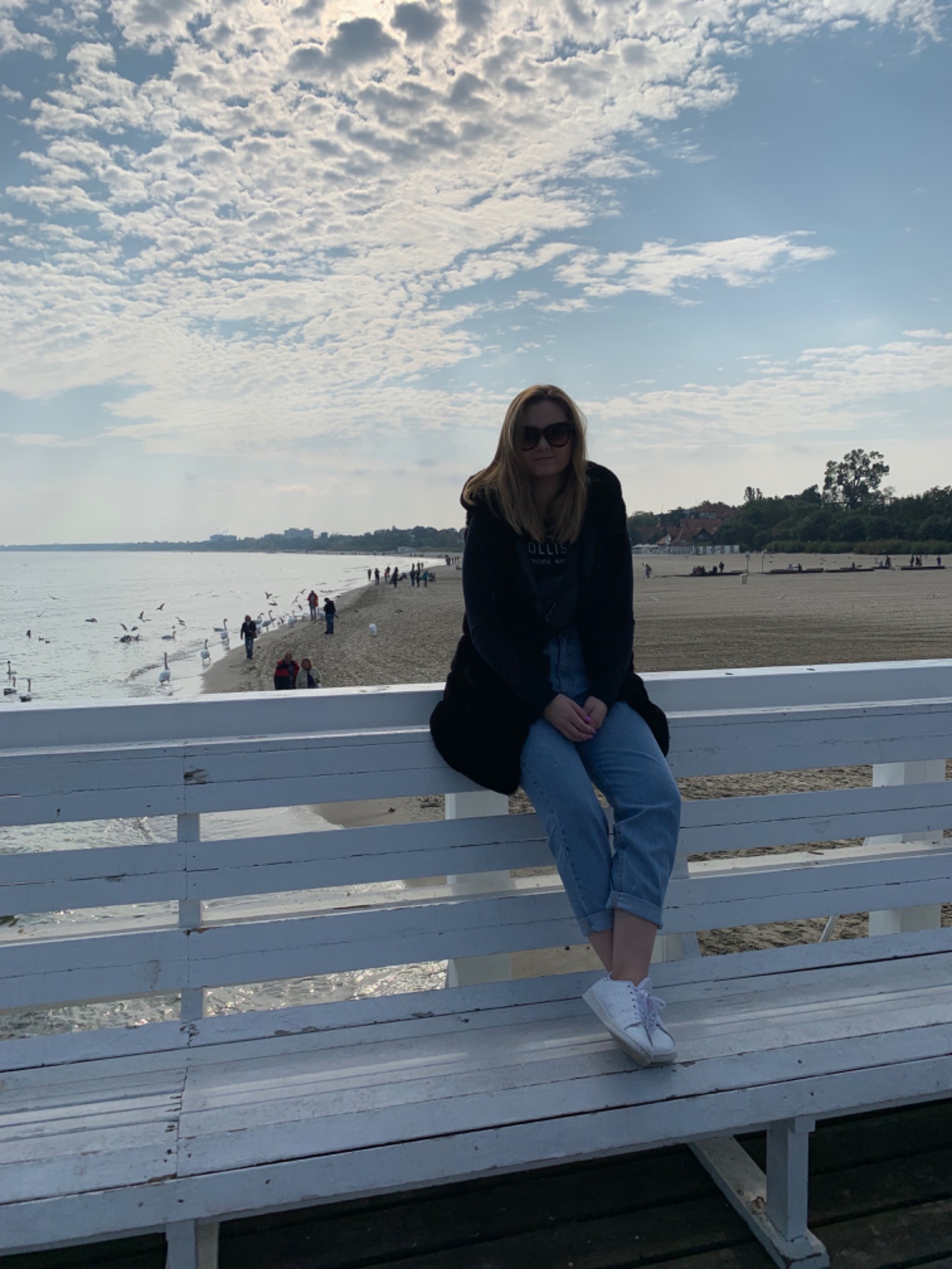 Kobieta w okularach przeciwsłonecznych siedzi na białej ławce na molo z widokiem na plażę i morze, gdzie widać spacerujących ludzi i pływające łabędzie.