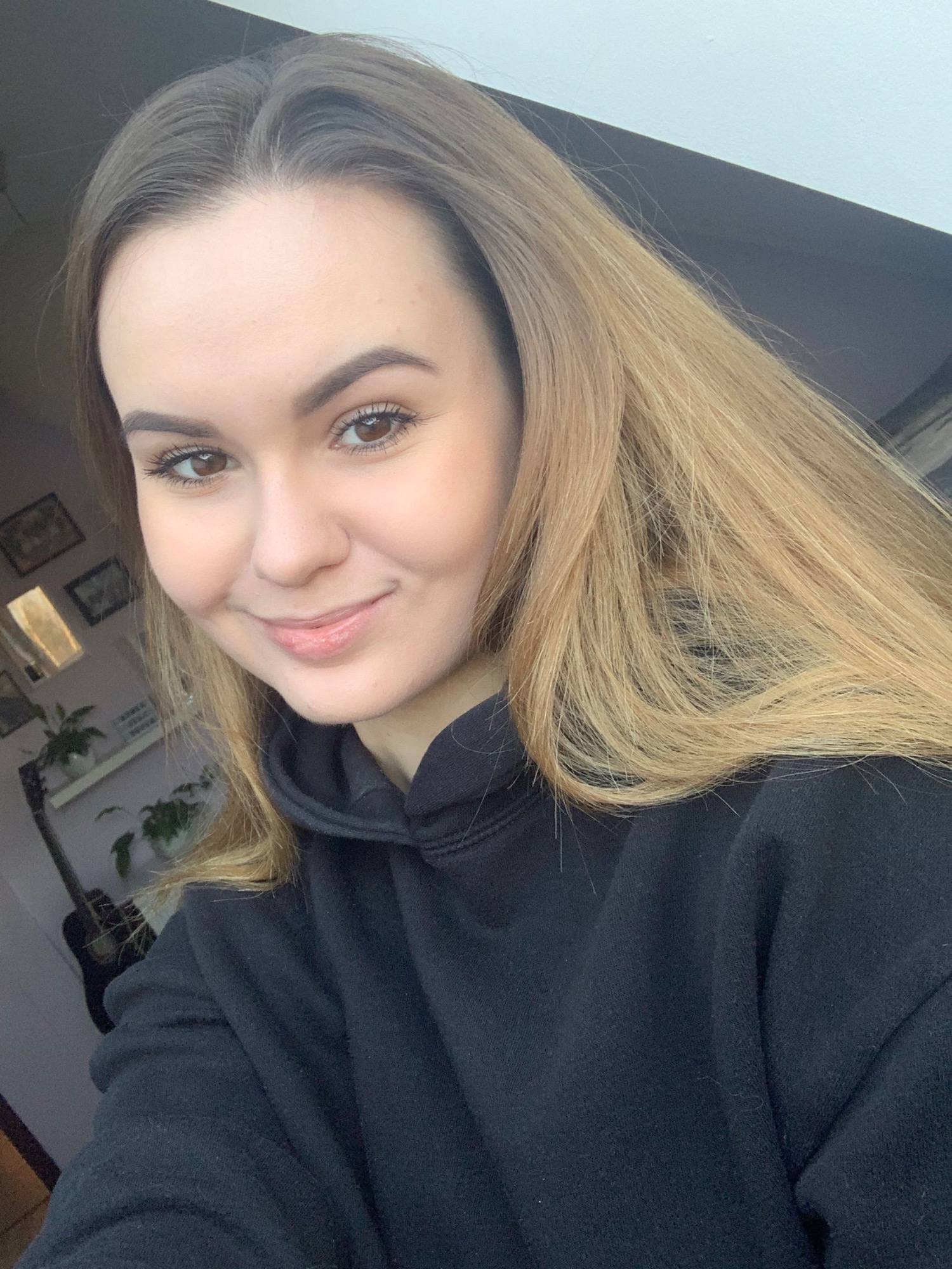 Portret uśmiechniętej młodej kobiety z długimi, ombre włosami w czarnej bluzie z kapturem, w tle półka z roślinami i gitara.