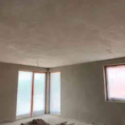 Pokój po nałożeniu świeżych, jasnoszarych gładzi, z zabezpieczonymi folią oknami i widoczną stertą piasku na podłodze.