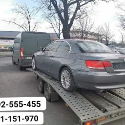 Szary samochód BMW 320i na lawecie w Warlubiu, widoczny również ciemny bus i inne pojazdy w tle.