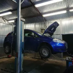 Niebieski samochód na podnośniku w warsztacie z otwartą maską, widoczne oświetlenie sufitowe i metalowe ściany.