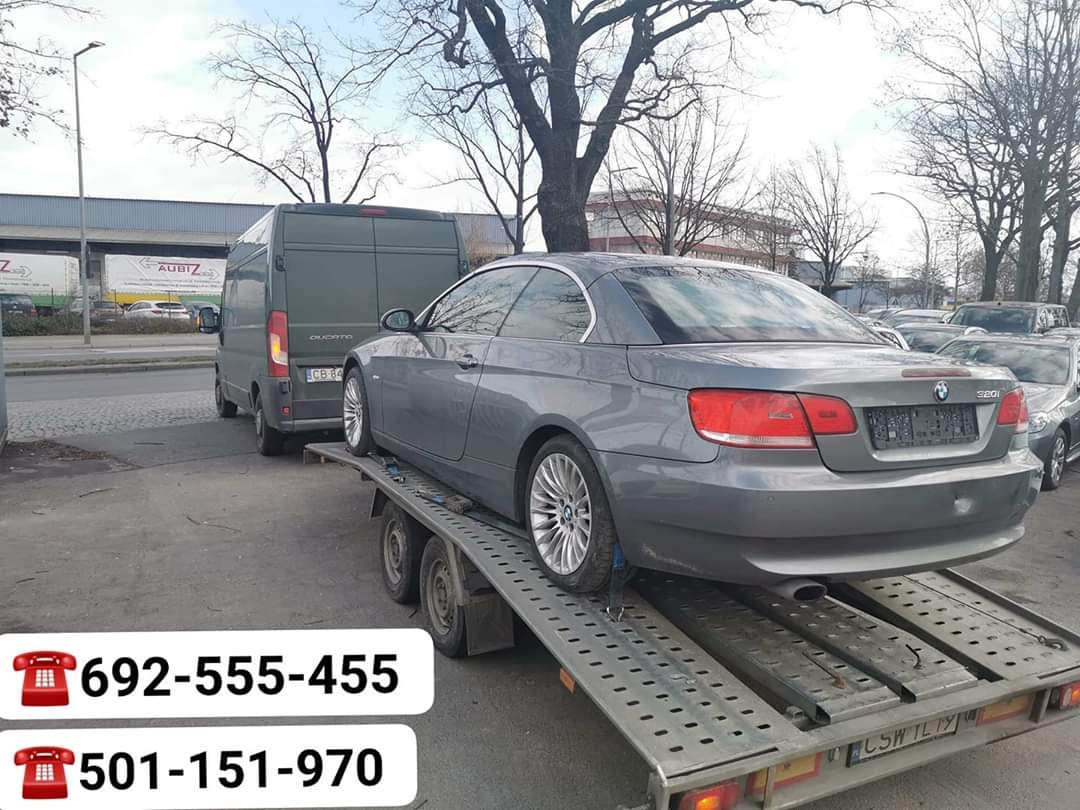 Szary samochód BMW 320i na lawecie w Warlubiu, widoczny również ciemny bus i inne pojazdy w tle.