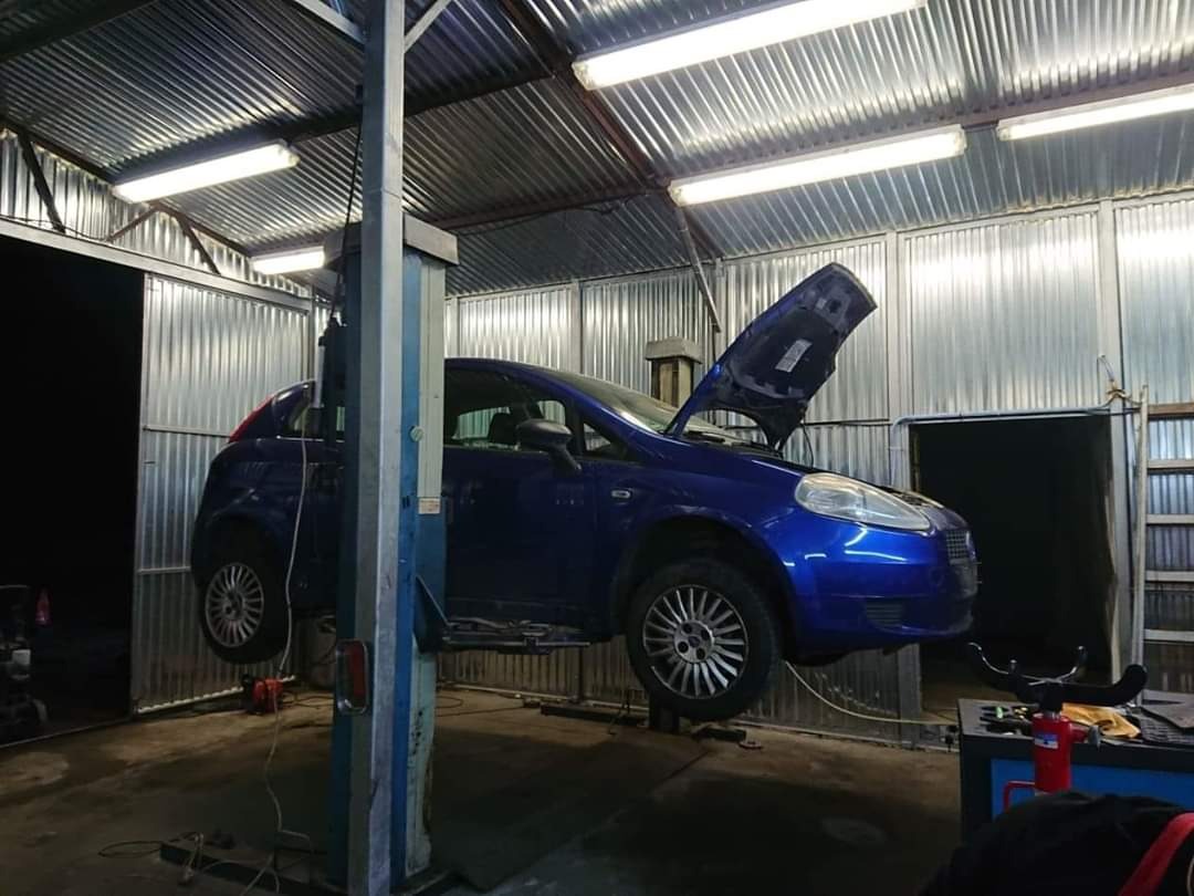 Niebieski samochód na podnośniku w warsztacie z otwartą maską, widoczne oświetlenie sufitowe i metalowe ściany.