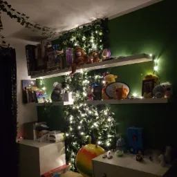 Pokój dziecięcy z zieloną ścianą, półkami ozdobionymi sztucznym bluszczem i lampkami, na których stoją zabawki i książki.