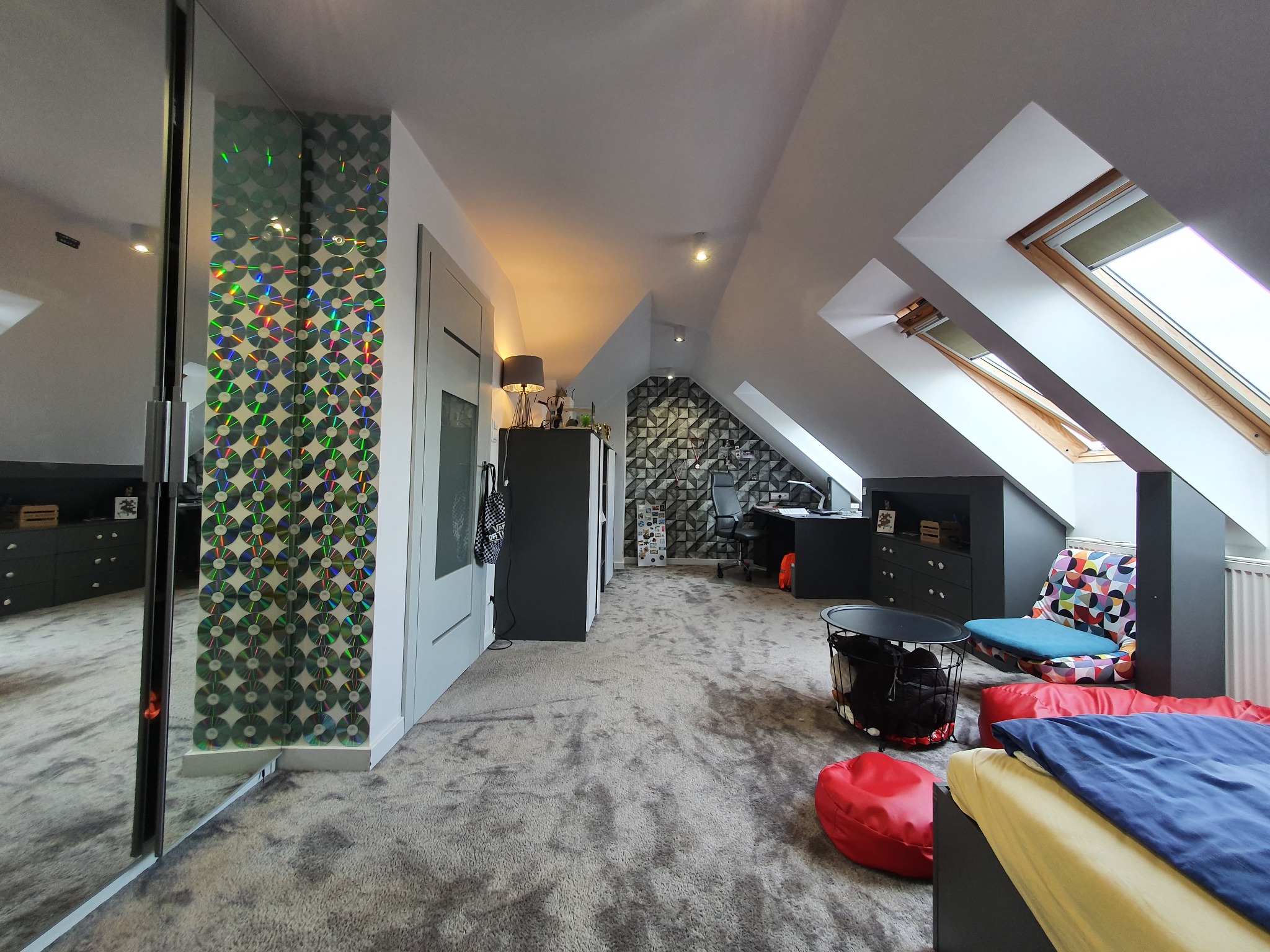 Pokój na poddaszu z oknami dachowymi, szarą wykładziną, szafą z lustrem ozdobioną płytami CD, biurkiem i miejscem do siedzenia z kolorowymi poduszkami.