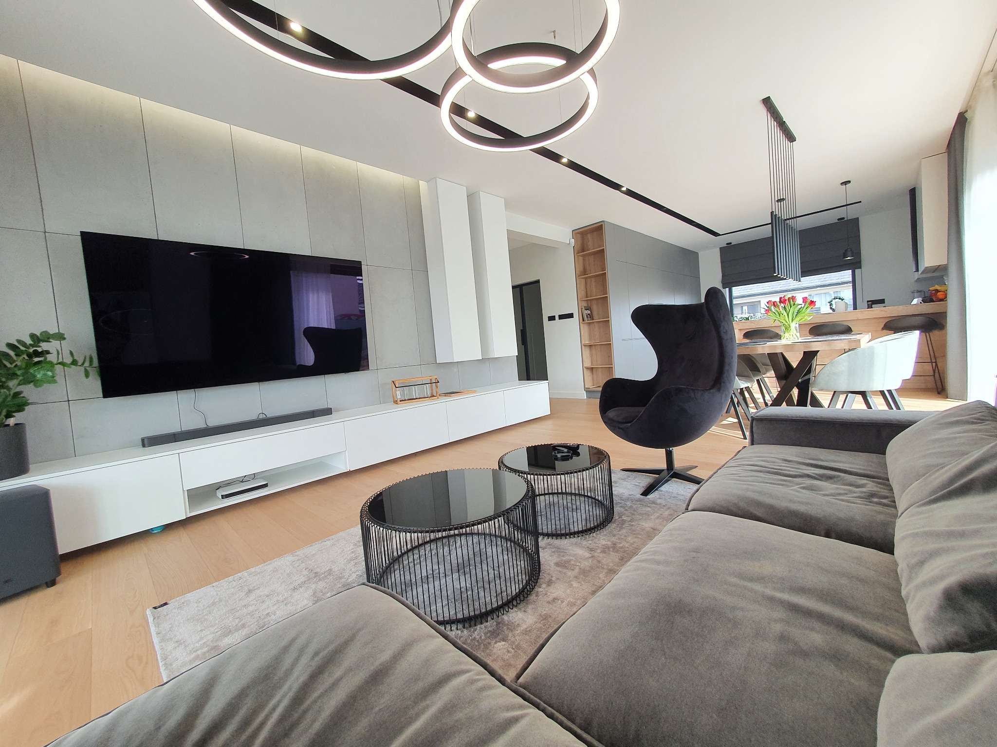 Nowoczesny salon z szarą sofą, okrągłymi stolikami kawowymi z czarnymi metalowymi podstawami, czarnym fotelem typu 'jajko' i podwieszanym oświetleniem w kształcie pierścieni.