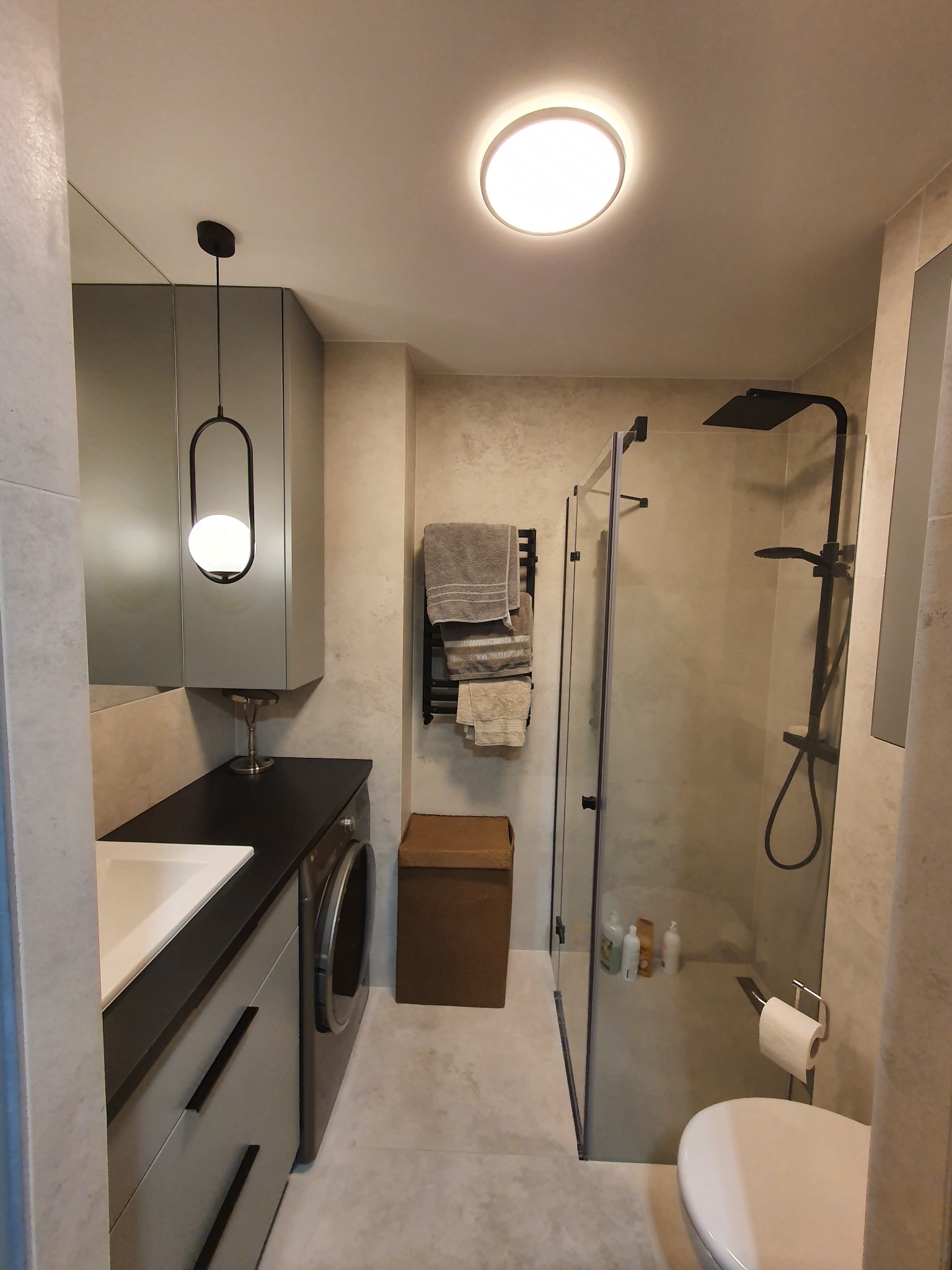 Nowoczesna łazienka z szarą zabudową, czarnym prysznicem typu deszczownica i pralką pod blatem, oświetlona lampą wiszącą z kulistym kloszem.