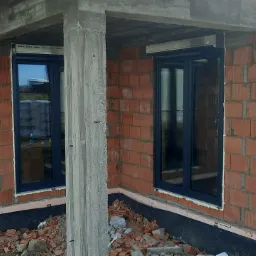 Surowy stan domu z czerwonej cegły z zamontowanymi oknami w ciemnych ramach, widoczne materiały budowlane i drabina.