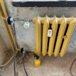 Żółty, żeliwny grzejnik z termostatem, podłączony do rur, w pomieszczeniu z łuszczącą się farbą na ścianach i zabrudzoną podłogą, obok butelka ze spryskiwaczem i narzędzie.