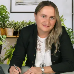 Kobieta w czarnej marynarce podpisuje dokument długopisem na biurku, w tle rośliny doniczkowe i półki z dekoracjami.