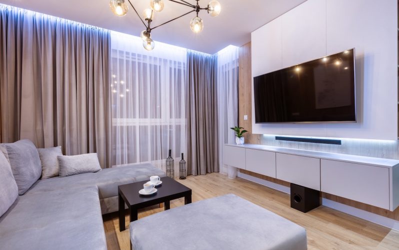 Nowoczesny salon z szarą sofą narożną, telewizorem na ścianie z podświetleniem LED, designerską lampą i drewnianą podłogą. Minimalistyczny wystrój.