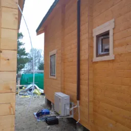 Zewnętrzna jednostka klimatyzacji zamontowana na drewnianej ścianie domu z widocznymi przewodami i narzędziami na ziemi, obok stoi zielony samochód dostawczy.