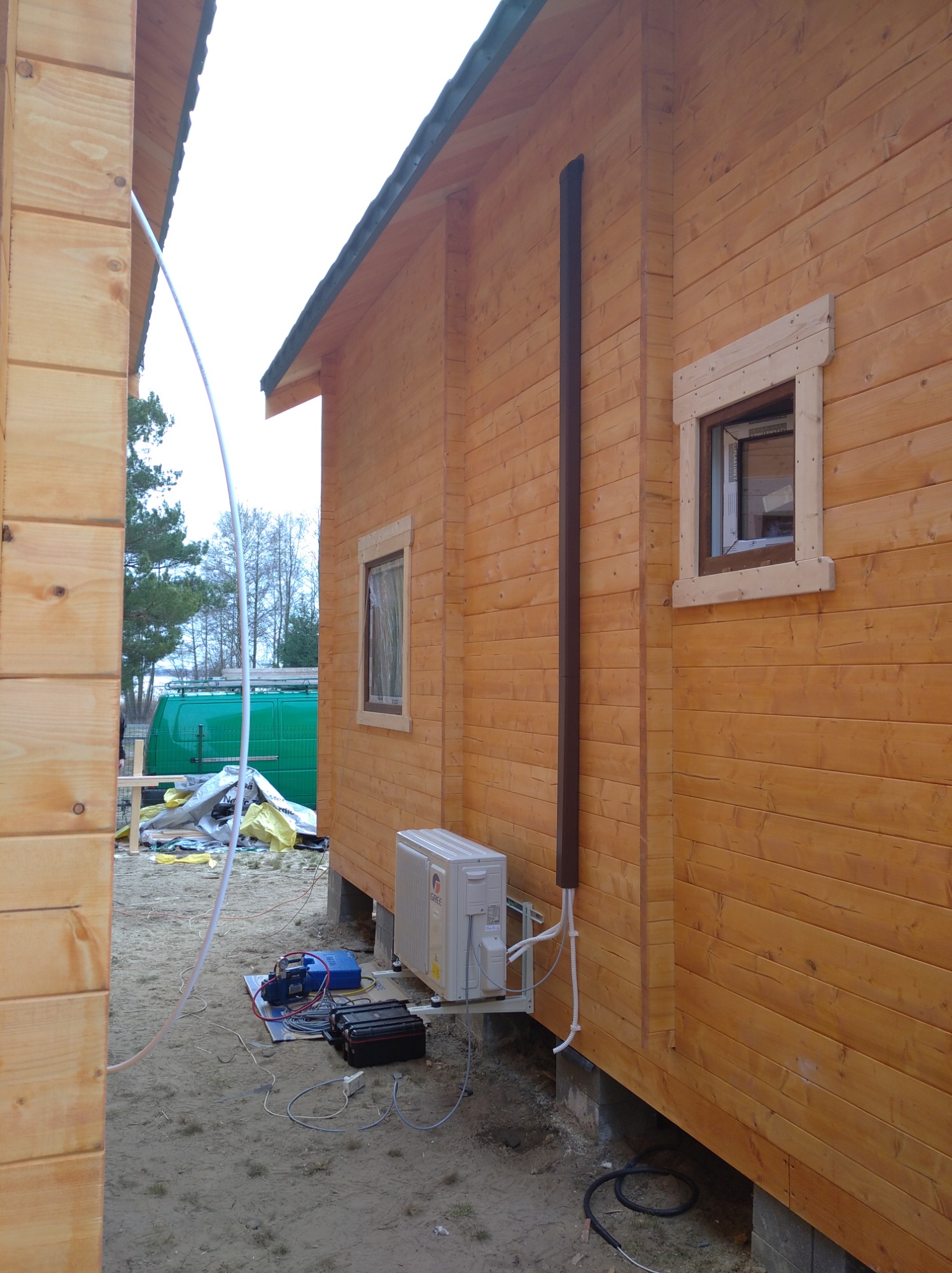 Zewnętrzna jednostka klimatyzacji zamontowana na drewnianej ścianie domu z widocznymi przewodami i narzędziami na ziemi, obok stoi zielony samochód dostawczy.