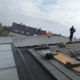 Dekarz na dachu z ciemnej dachówki, przenosi płytki, widoczne narzędzia i akcesoria dekarskie, w tle zabudowa miejska i pochmurne niebo.