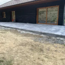 Nowoczesny taras z szarych płyt kamiennych w trakcie budowy, widoczny zagęszczarka wibracyjna, dom w stanie surowym zamkniętym w tle.