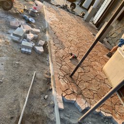 Patryk Pęcak - Układanie nieregularnej kostki brukowej w odcieniach brązu i beżu na podjeździe, widoczne narzędzia pracy, wózek, stos kostki, obrzeża i częściowo ułożona nawierzchnia.