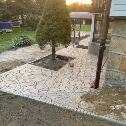 Patryk Pęcak - Kremowa, nieregularna nawierzchnia z kamienia ułożona wokół drzewa iglastego, obrzeża z szarej kostki, widoczny fragment elewacji domu z kamienną okładziną, w tle zabudowa i zachodzące słońce.