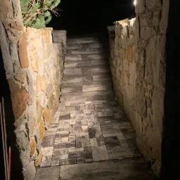 Kamienne schody prowadzące w górę, oświetlone punktowym światłem, otoczone murem z nieregularnych kamieni, z fragmentem iglastego drzewa w rogu.