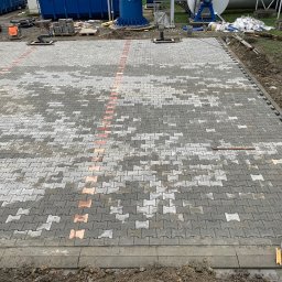 Patryk Pęcak - Ułożona szara kostka brukowa typu 'psia kość' z pomarańczowymi akcentami, widoczne studzienki kanalizacyjne i zaparkowany samochód w tle, teren budowy.