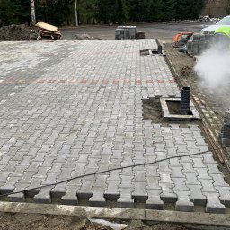 Patryk Pęcak - Fragment placu z ułożoną szarą kostką brukową typu I, widoczny pracownik w odblaskowej kamizelce, palety z kostką w tle, oraz rury kanalizacyjne w przygotowanych otworach.