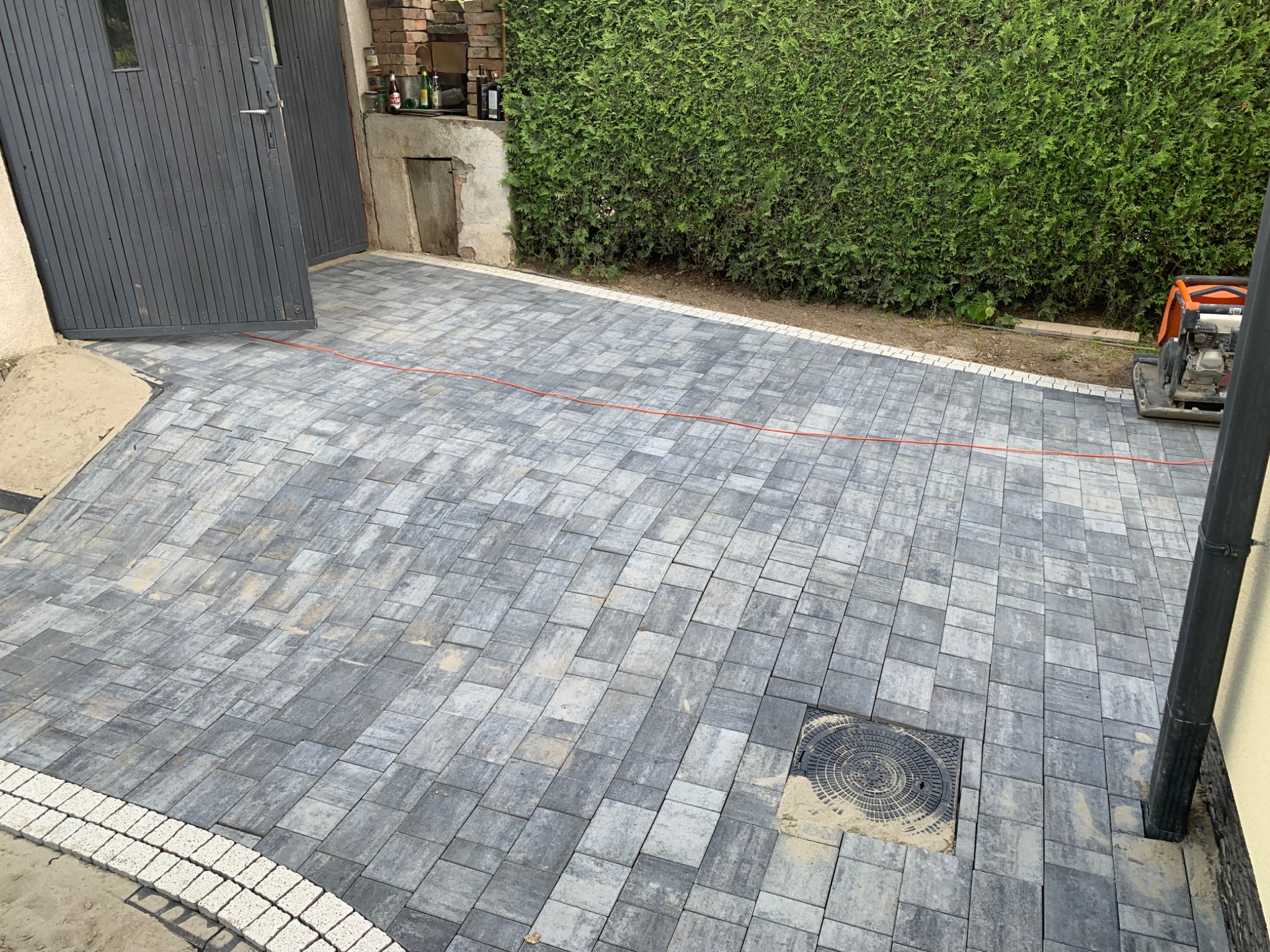 Ułożona szara kostka brukowa z białym obramowaniem na podwórku w Żyznowie. Widoczny właz kanalizacyjny i ubijarka w tle, oraz zielony żywopłot.