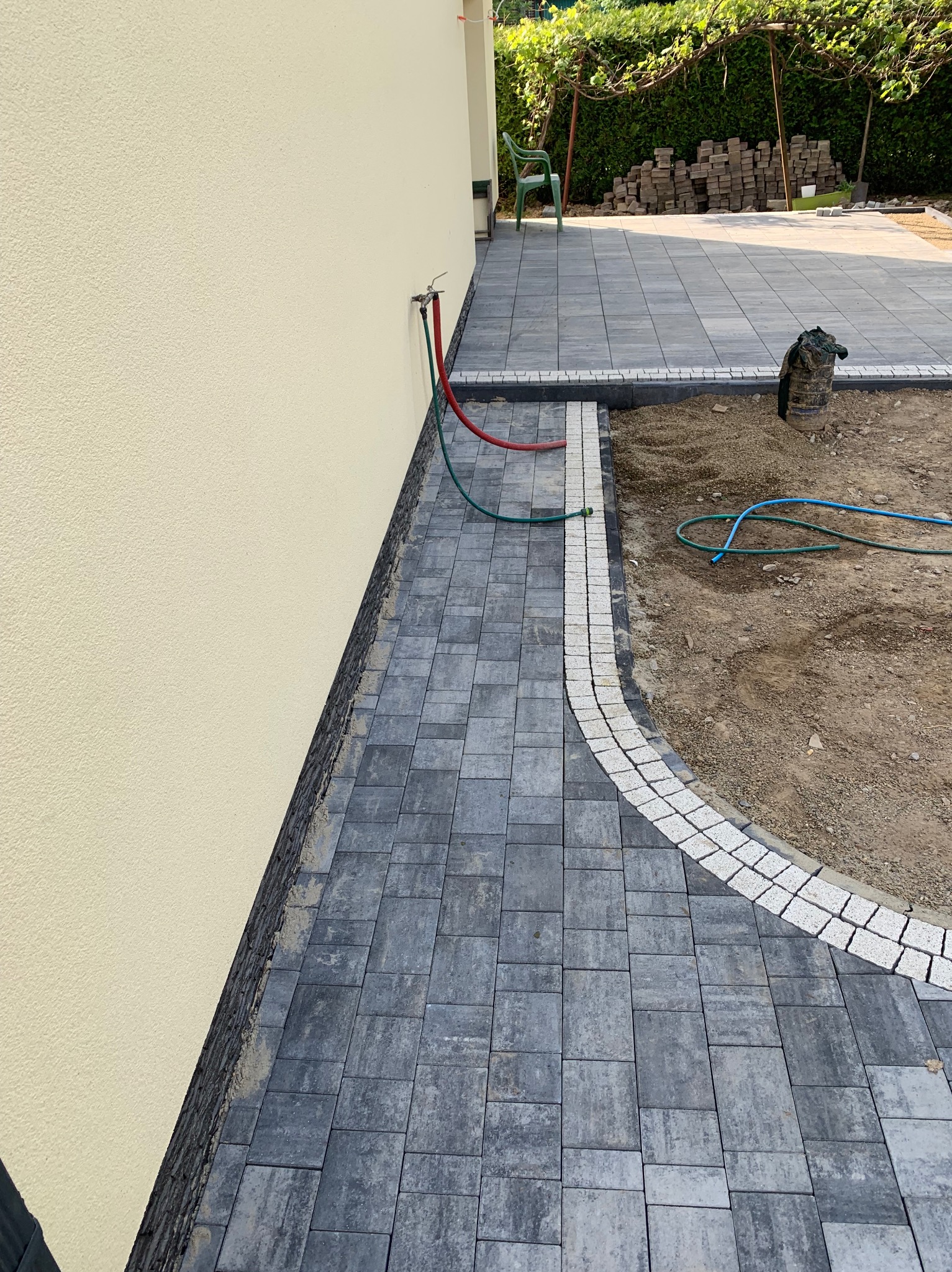 Częściowo ułożony chodnik z szarych kostek brukowych z białym obramowaniem, widoczny fragment budynku z beżową elewacją, węże ogrodowe i przygotowany teren pod dalsze prace.