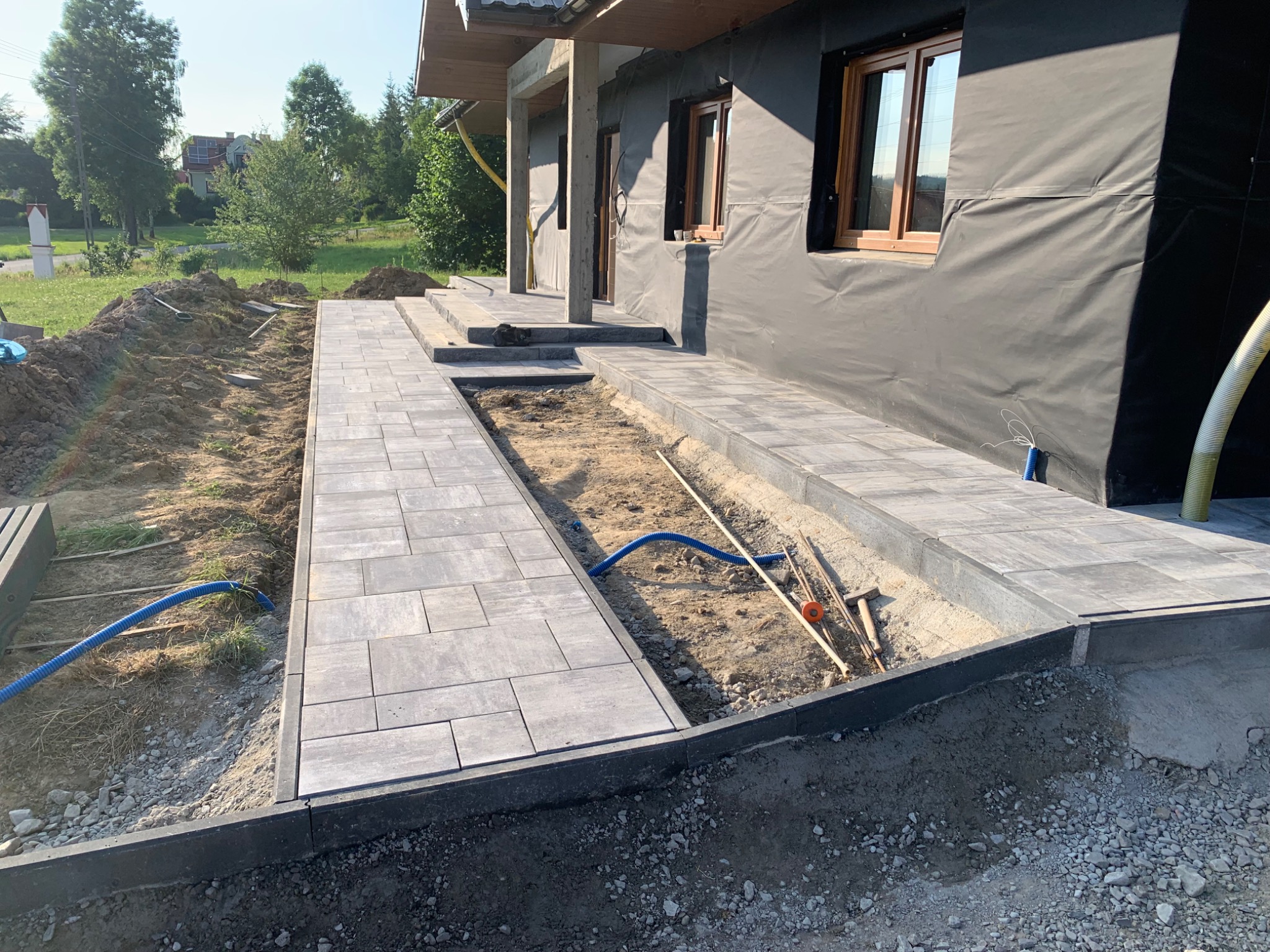 Częściowo ułożony chodnik z szarych płyt betonowych obok domu z czarną folią izolacyjną na ścianach, widoczne narzędzia i niebieski wąż na ziemi.