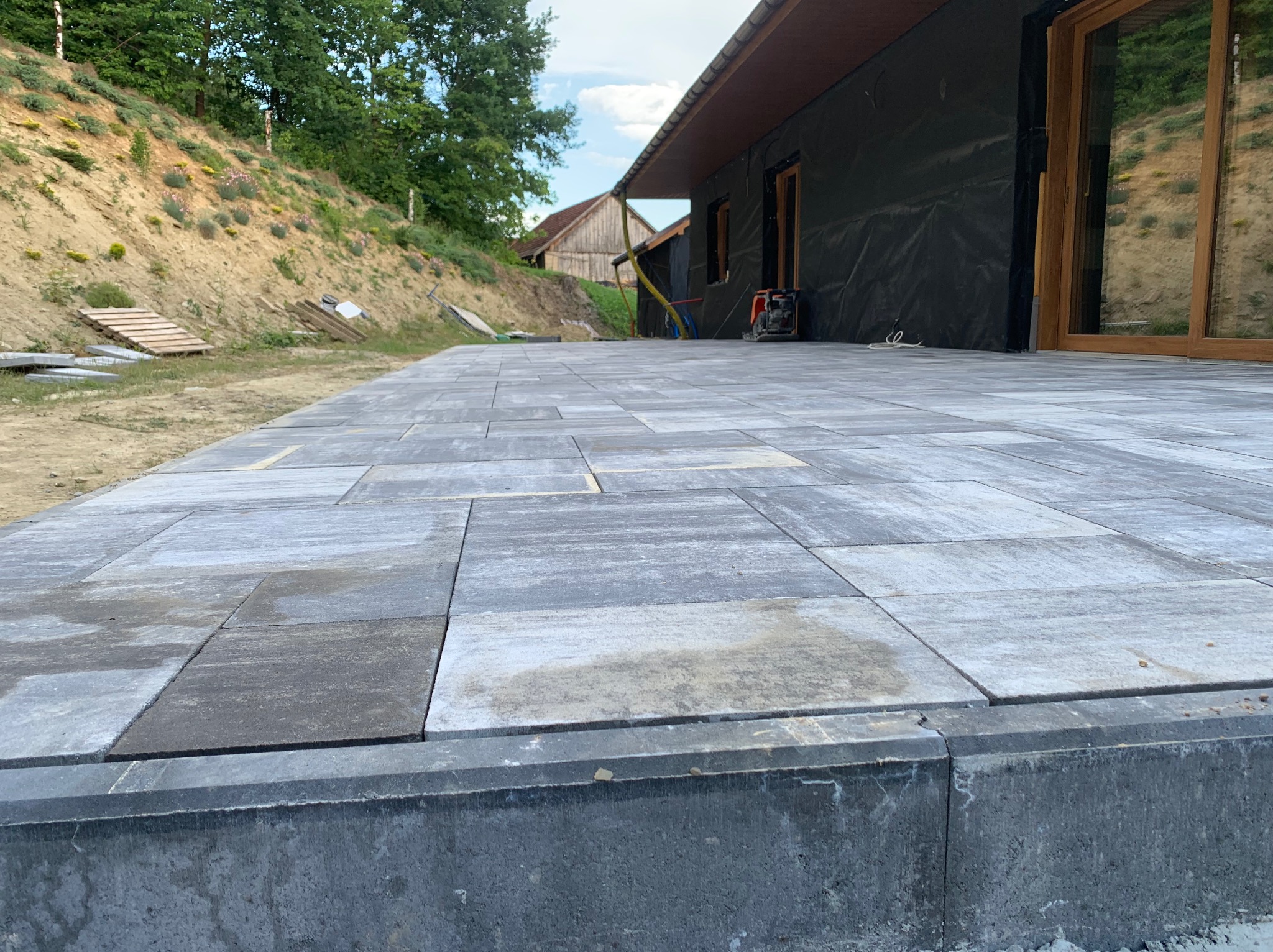 Szary taras z płyt betonowych z widocznym fragmentem budynku w trakcie wykończenia i ziemnym nasypem w tle.